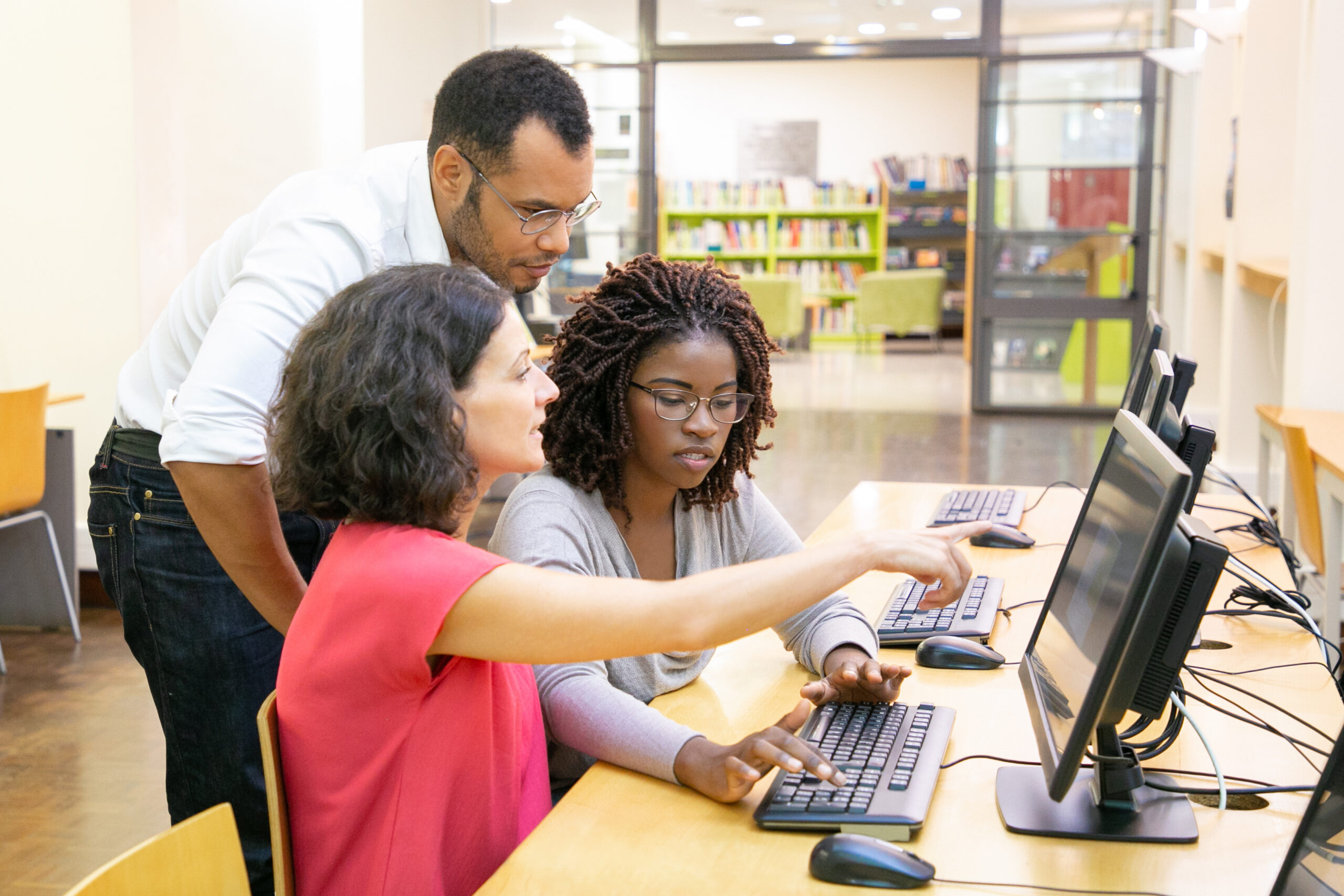 Instructor helping students in computer class. Man and women sitting and standing at desk, using desktop, pointing at monitor and talking.