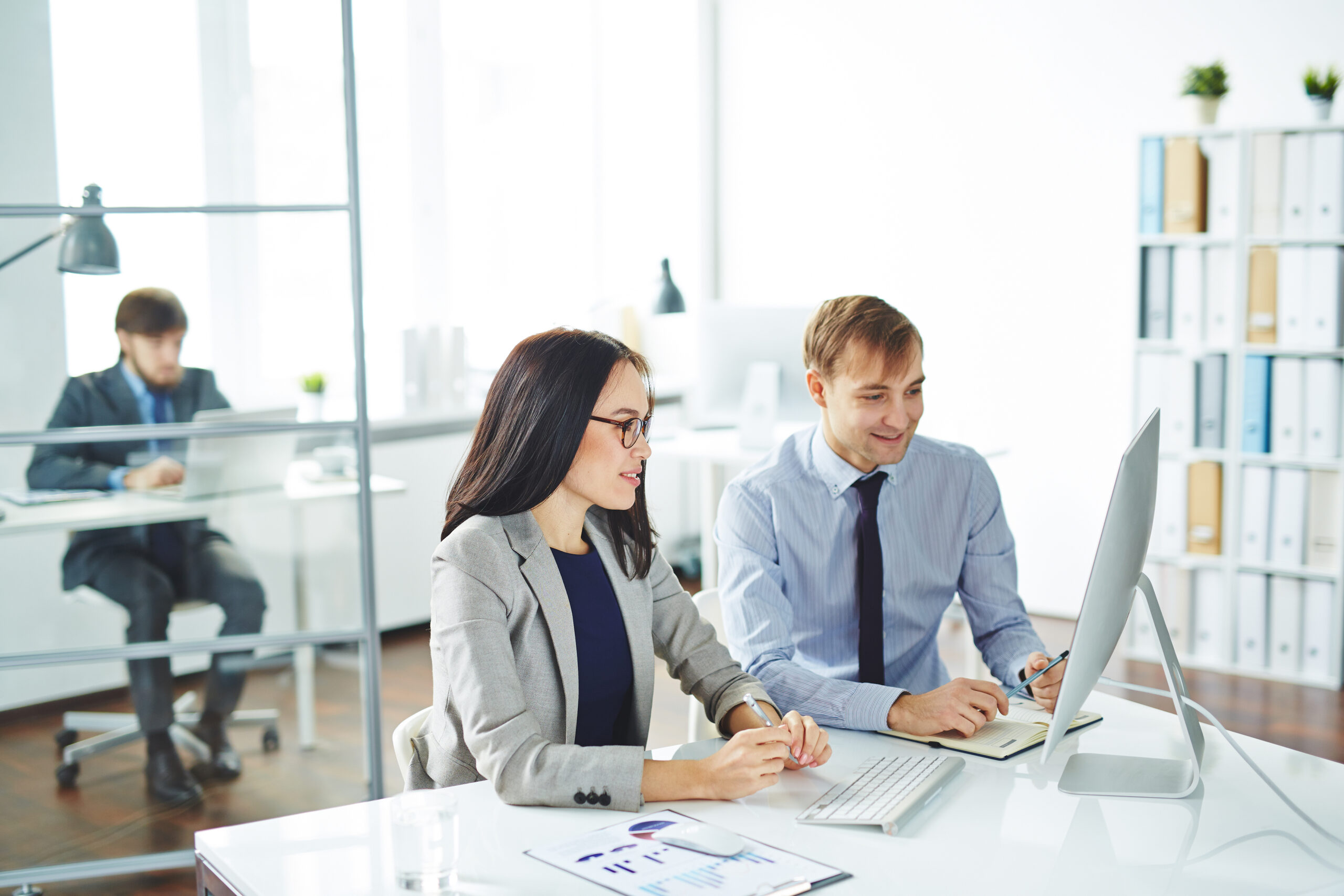 Two office professionals reviewing data on a computer together in a bright, modern workspace with charts and documents on the desk.