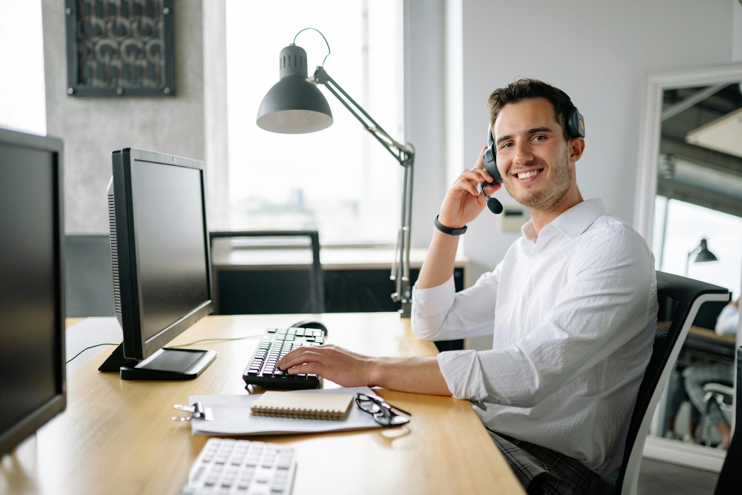 Smiling IT support specialist wearing a headset, sitting at a desk with a computer and office supplies in a bright modern workspace.