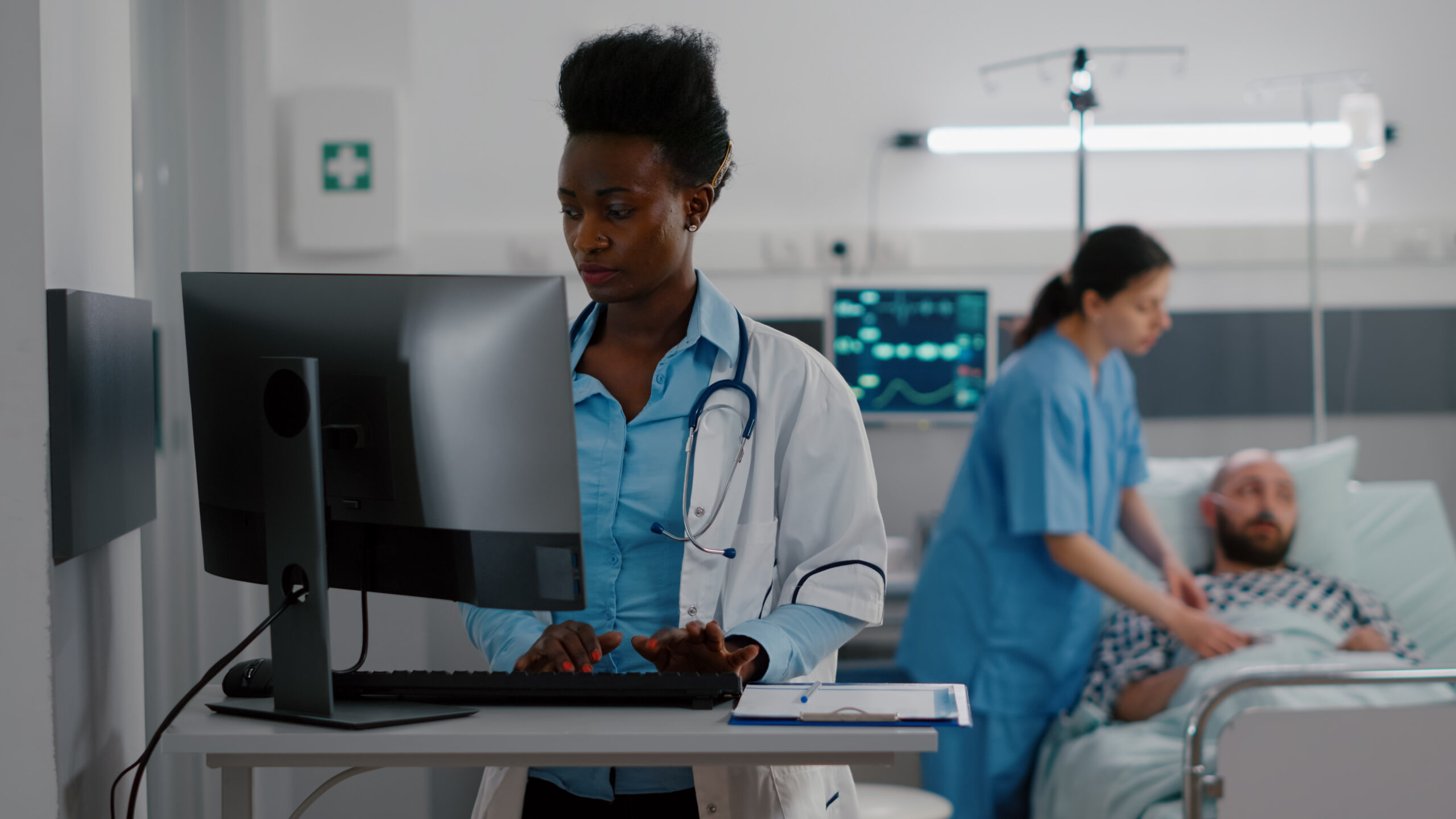 Doctor typing on computer while nurse checks on patient in the background Doctor typing on computer while nurse checks on patient in the background